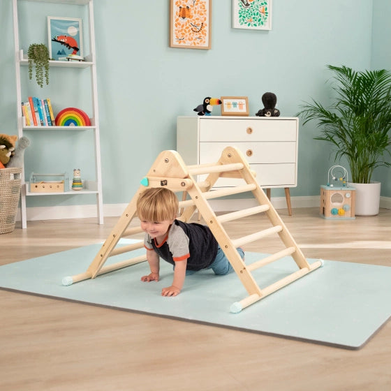 Blonder kleiner Junge krabbelt im Kinderzimmer unter Pikler Dreieck TP Toys hindurch und lacht